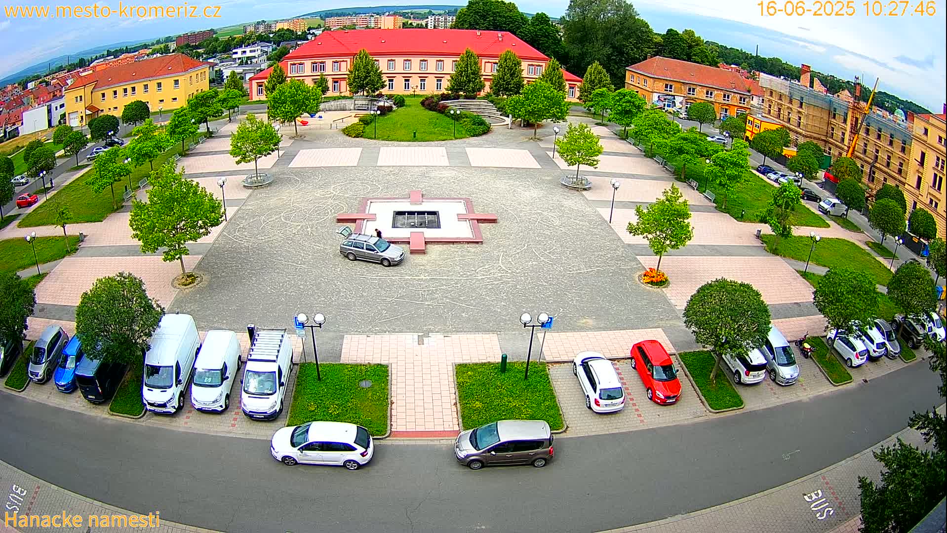 An aerial view of a town square with parked cars, surrounded by buildings and lush green landscaping under partly cloudy skies.