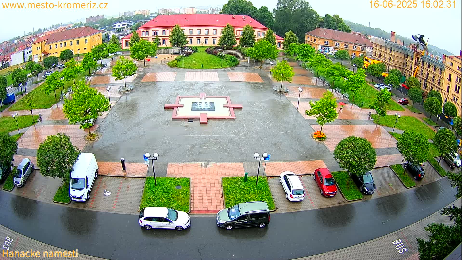 A wet town square with a fountain in the center, surrounded by parked cars and lush green trees under a rainy sky.