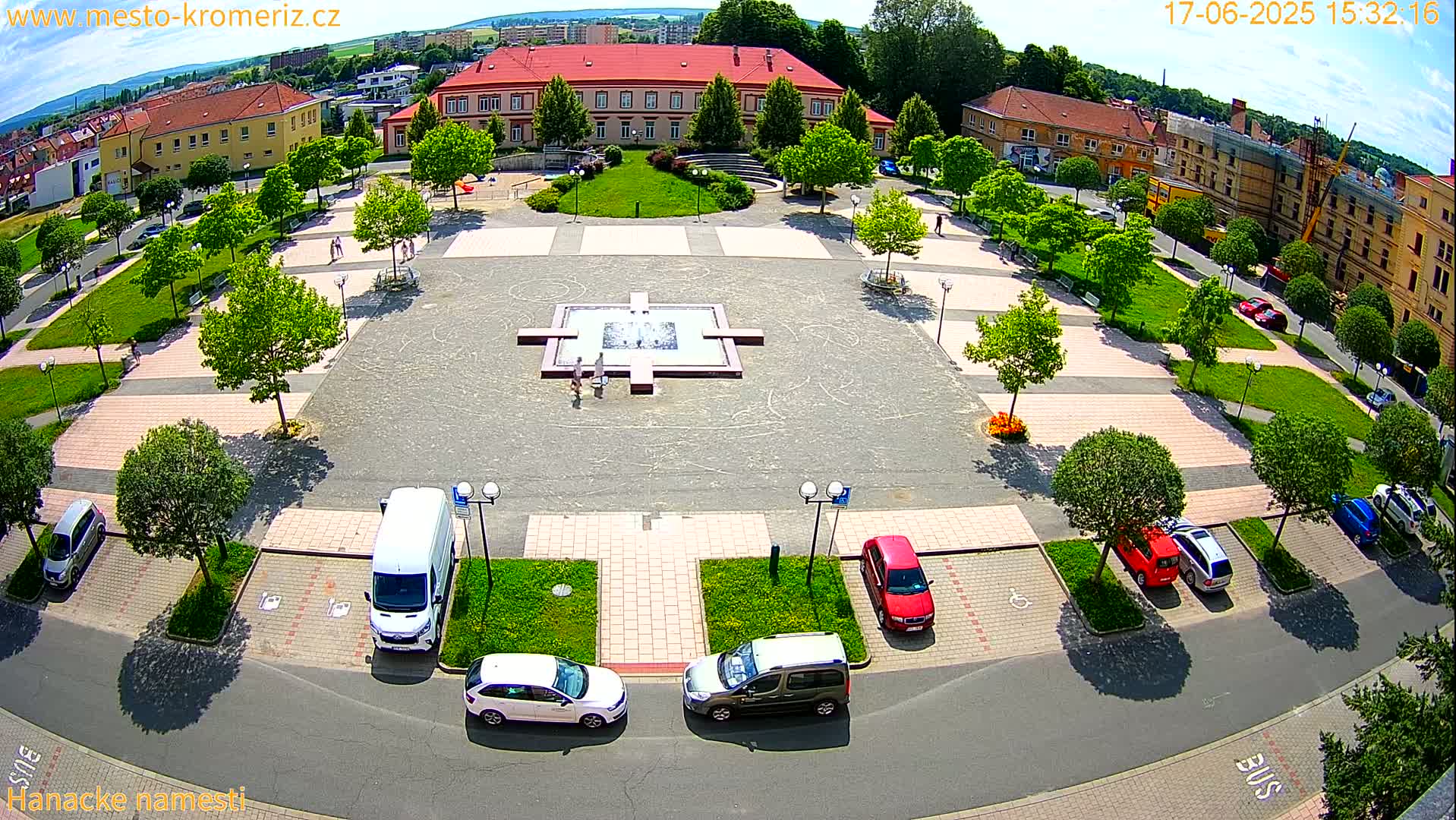 An aerial view of a town square on a sunny day shows a central fountain, surrounding green spaces, parked cars, and several buildings in the background.