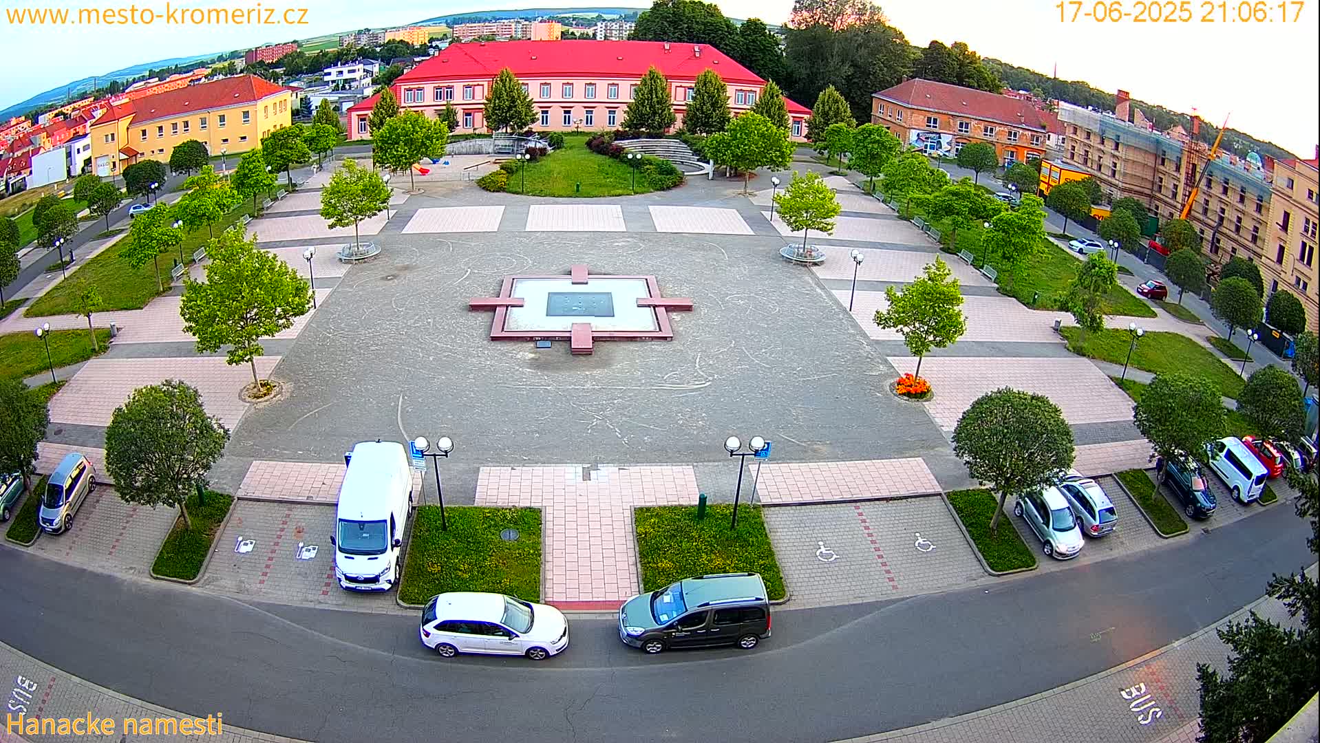 An aerial view of a town square at dusk, featuring a central fountain, several parked cars, and buildings surrounding the paved area with landscaping and trees.