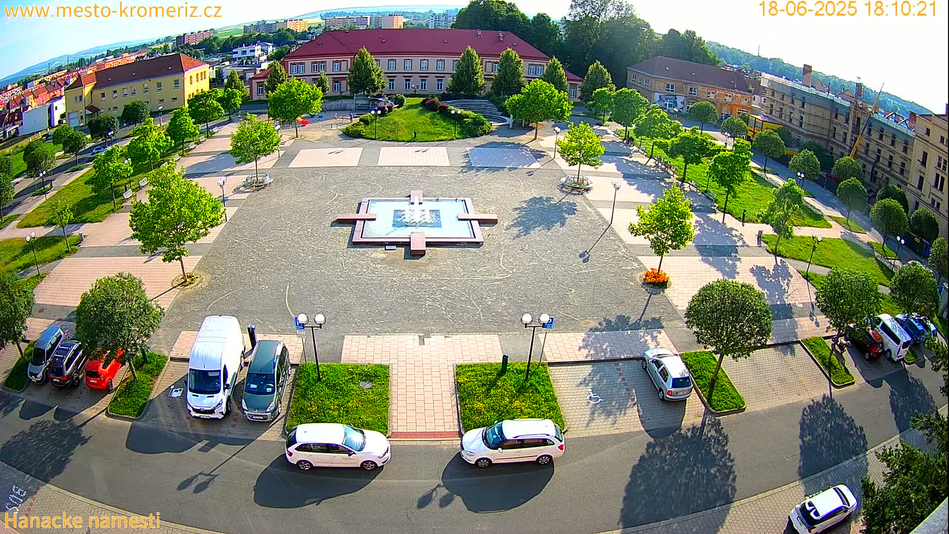An aerial view of a town square on a sunny day, showing a central fountain, surrounding green spaces, parked cars, and buildings in the background.