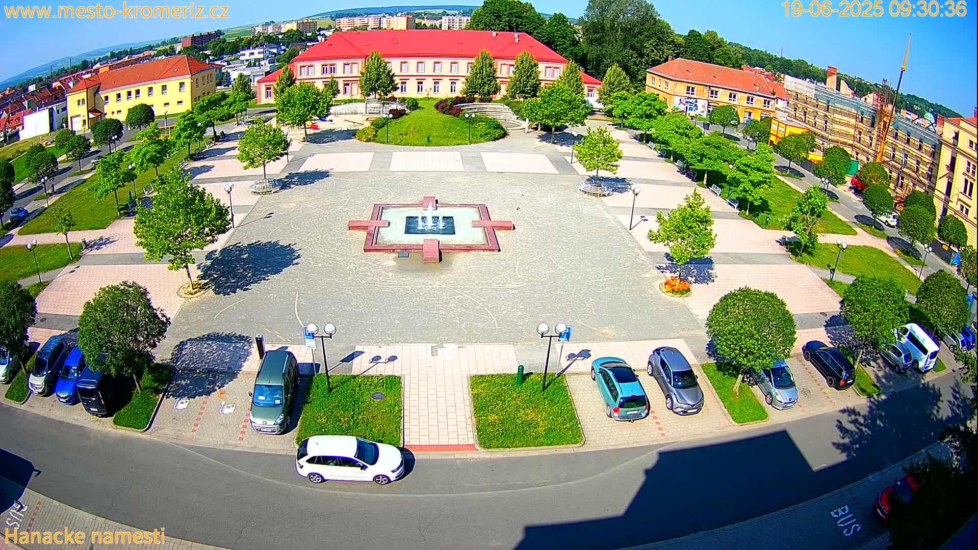 An aerial view of a town square on a sunny day, featuring a central fountain, surrounding buildings, and cars parked along the edges.