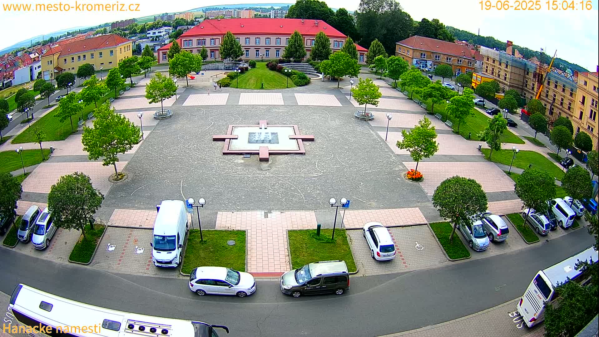 An aerial view of a town square on a partly sunny day, showing a central fountain, surrounding parked cars, and buildings in the background.