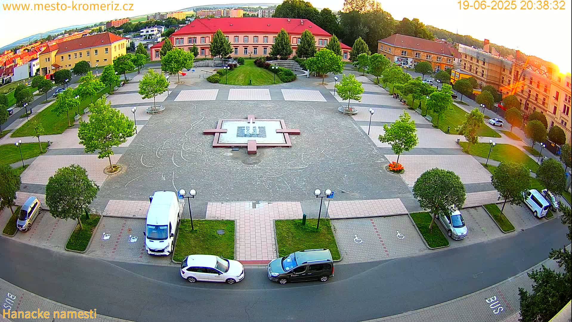 An aerial view of a town square at sunset, showing a central plaza with a fountain, surrounded by parked cars, buildings, and green spaces.