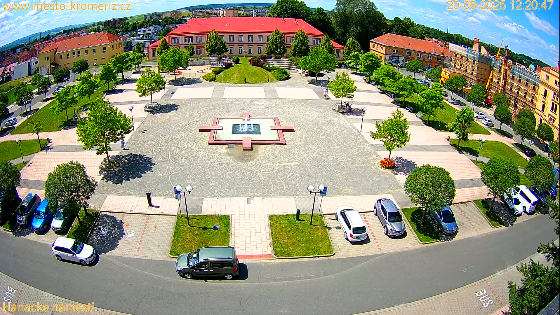 An aerial view of a town square with a fountain in the center, surrounded by buildings, trees, and parked cars on a sunny day.