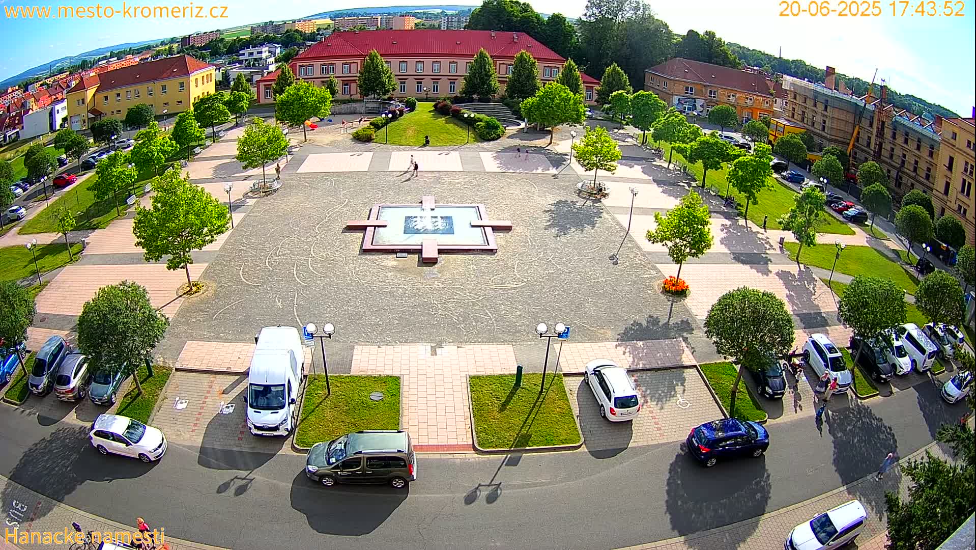 An aerial view of a town square on a sunny day, showing a fountain at the center, surrounded by cars parked along the streets and green spaces with trees.