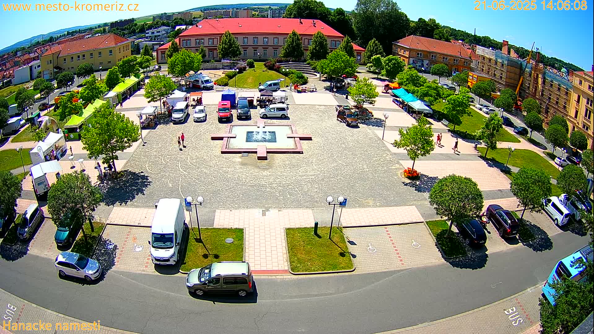 An aerial view of a town square on a sunny day, showing a market with several stalls and vehicles parked around a central fountain.