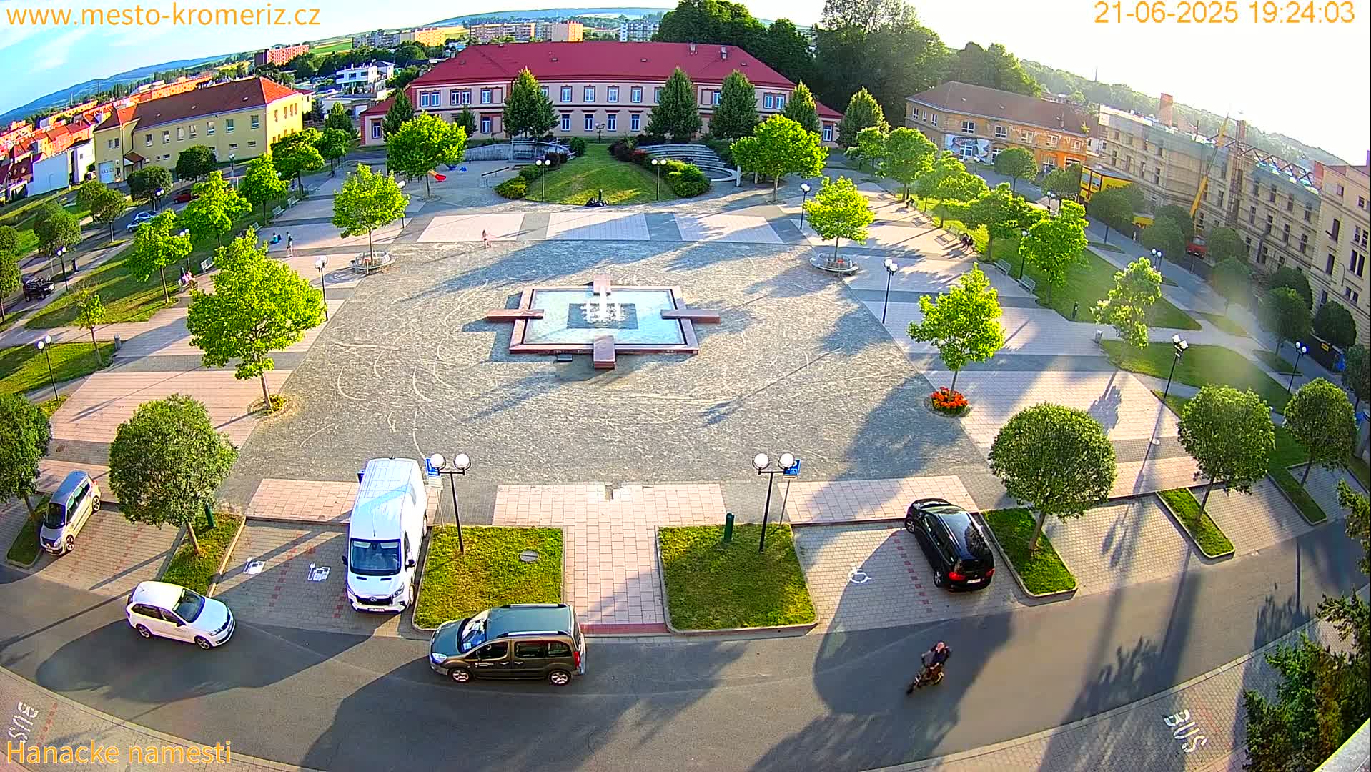 An aerial view shows a town square with a fountain at its center, surrounded by trees, buildings, and parked cars, on a sunny day.