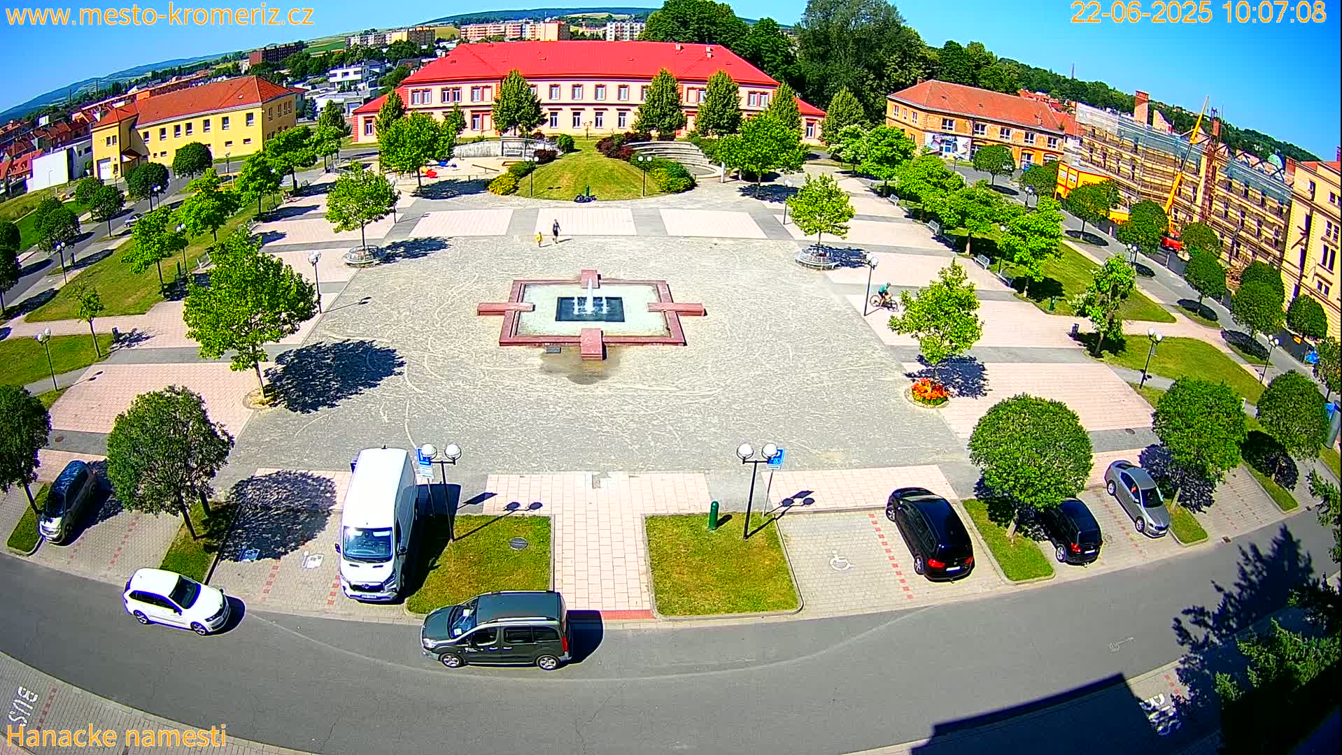 A town square with a fountain at its center, surrounded by parked cars and buildings, on a sunny day.