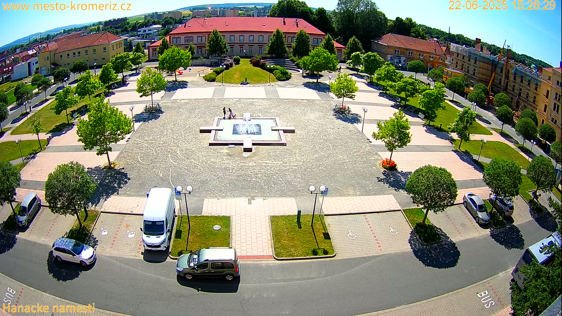 An aerial view of a town square with a fountain, surrounded by parked cars, green spaces, and buildings under sunny conditions.