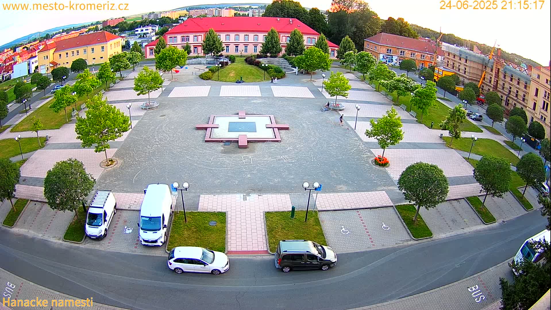 An aerial view of a town square at dusk, showing a paved area with a central fountain, surrounding buildings, parked cars, and landscaping with green trees.