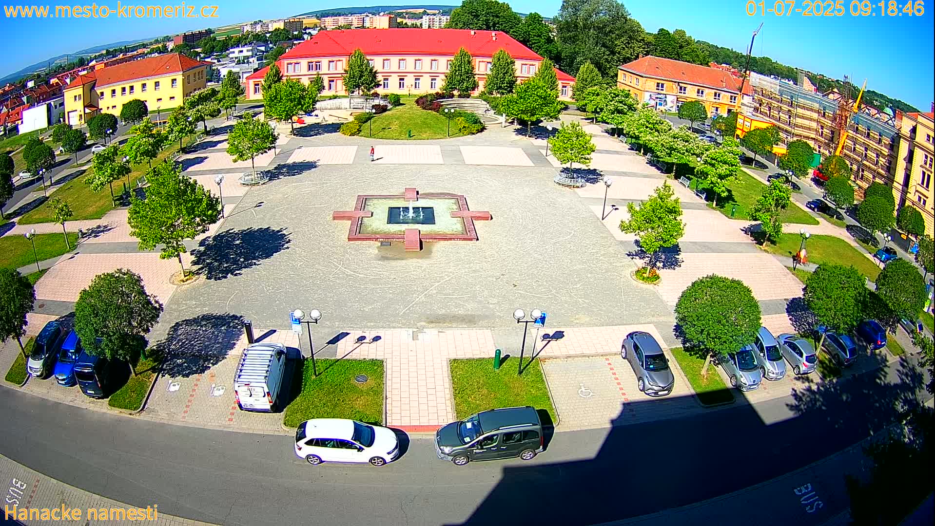A sunny day reveals a town square with a central fountain, surrounded by parked cars and buildings, including a long red building.