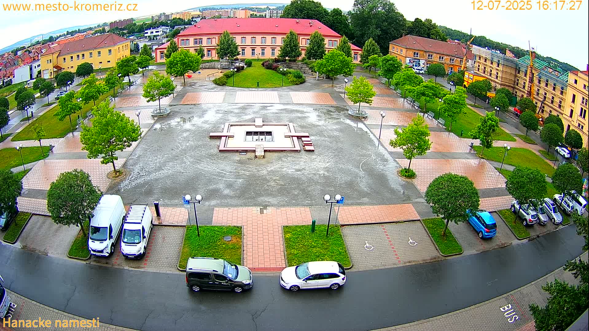 A sunny day reveals a town square with a central fountain, surrounded by parked cars and buildings, including a long red building.