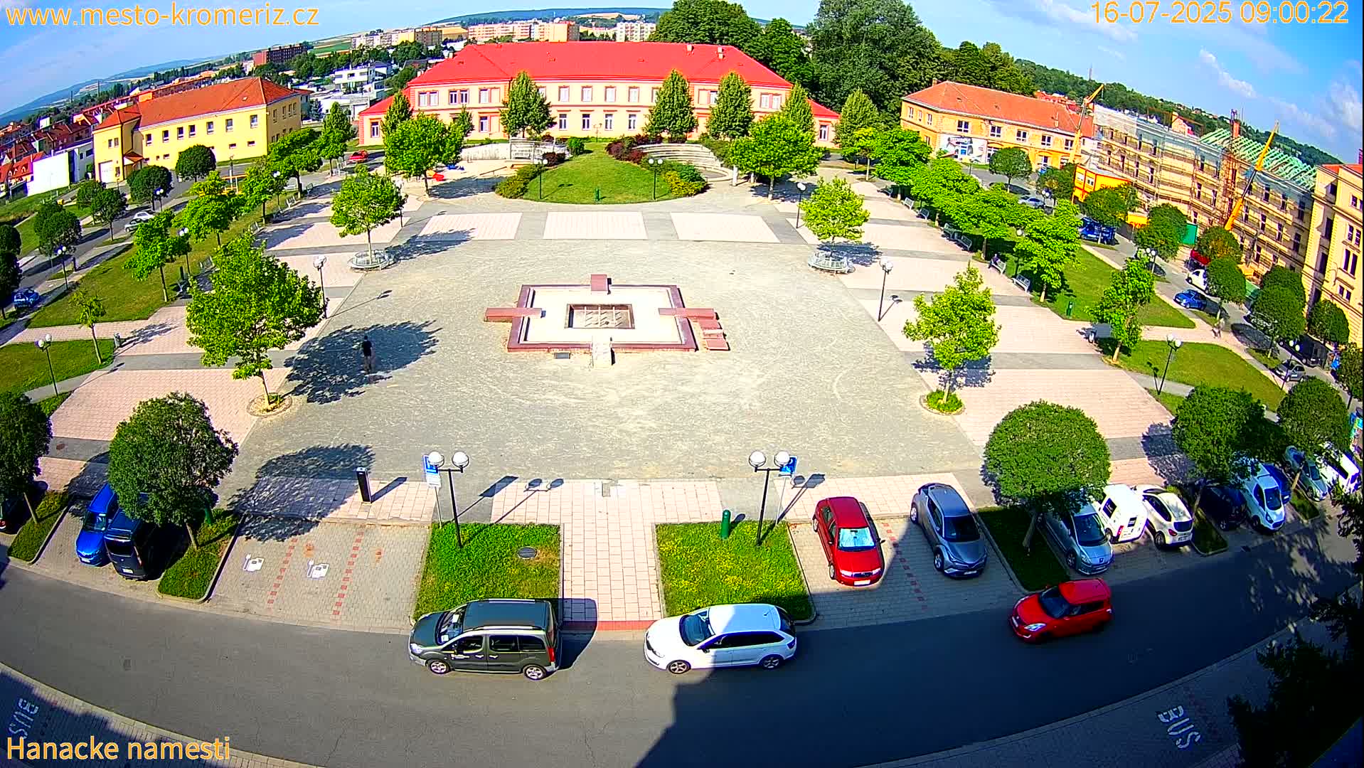 A town square with a central fountain, surrounded by buildings, trees, and parked cars under sunny conditions.