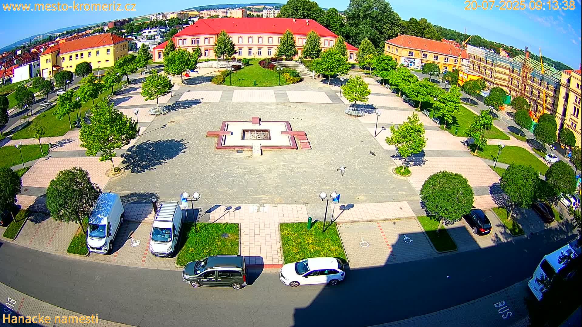 A town square with a central fountain, surrounded by buildings, green spaces, and parked cars under a sunny sky.