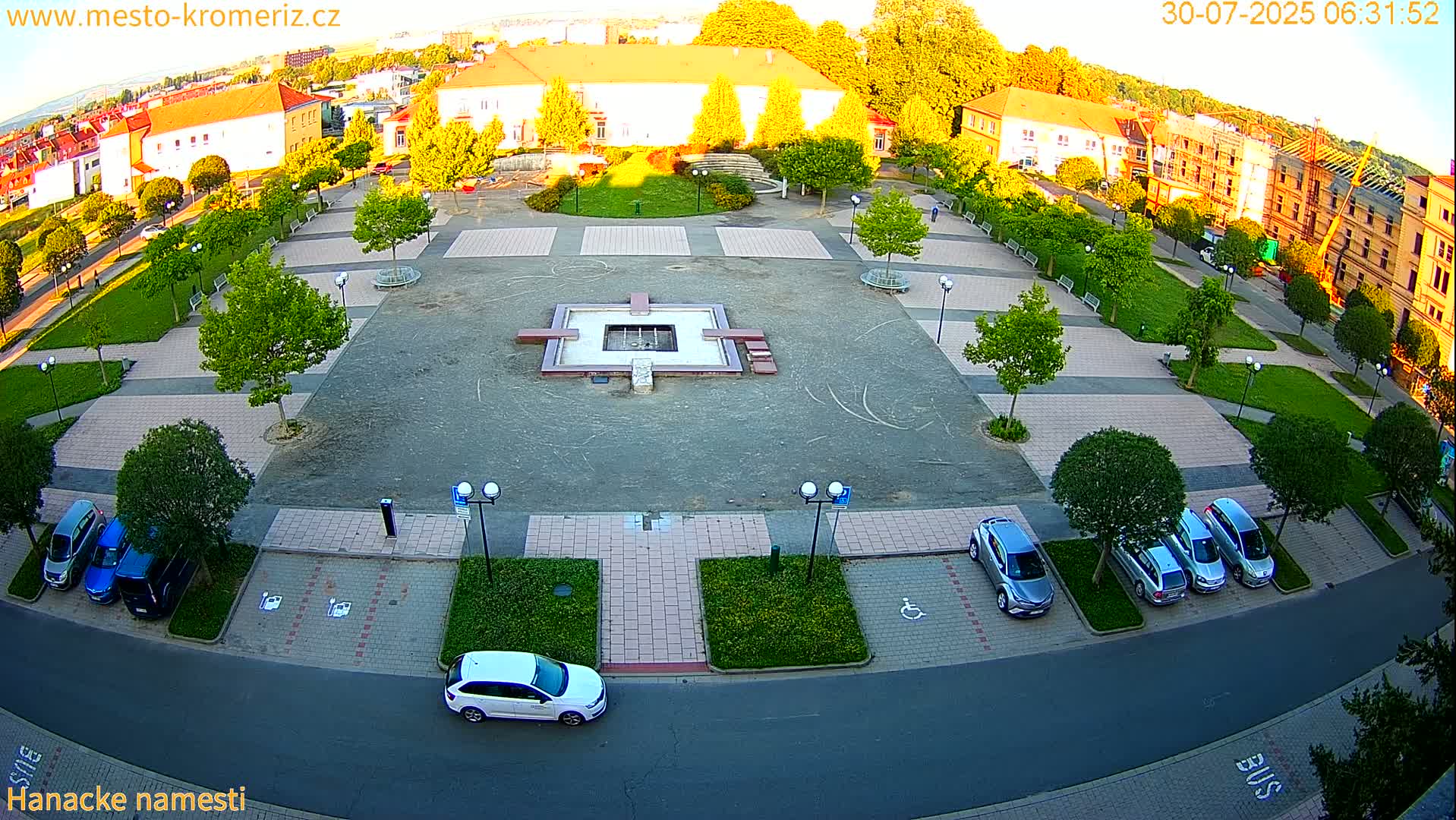A town square with a central fountain, surrounded by buildings, green spaces, and parked cars under a sunny sky.
