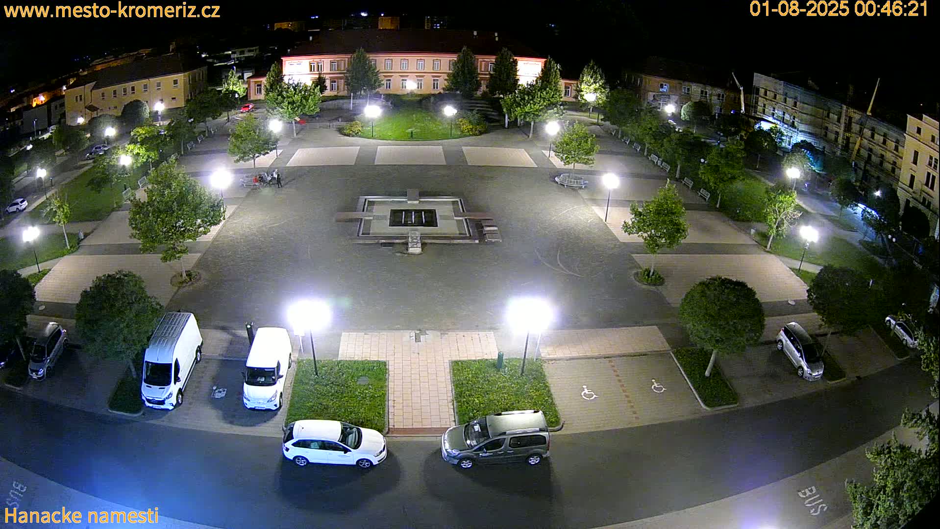 A nighttime, overhead view of a town square shows a central fountain, surrounding landscaping, and parked cars.
