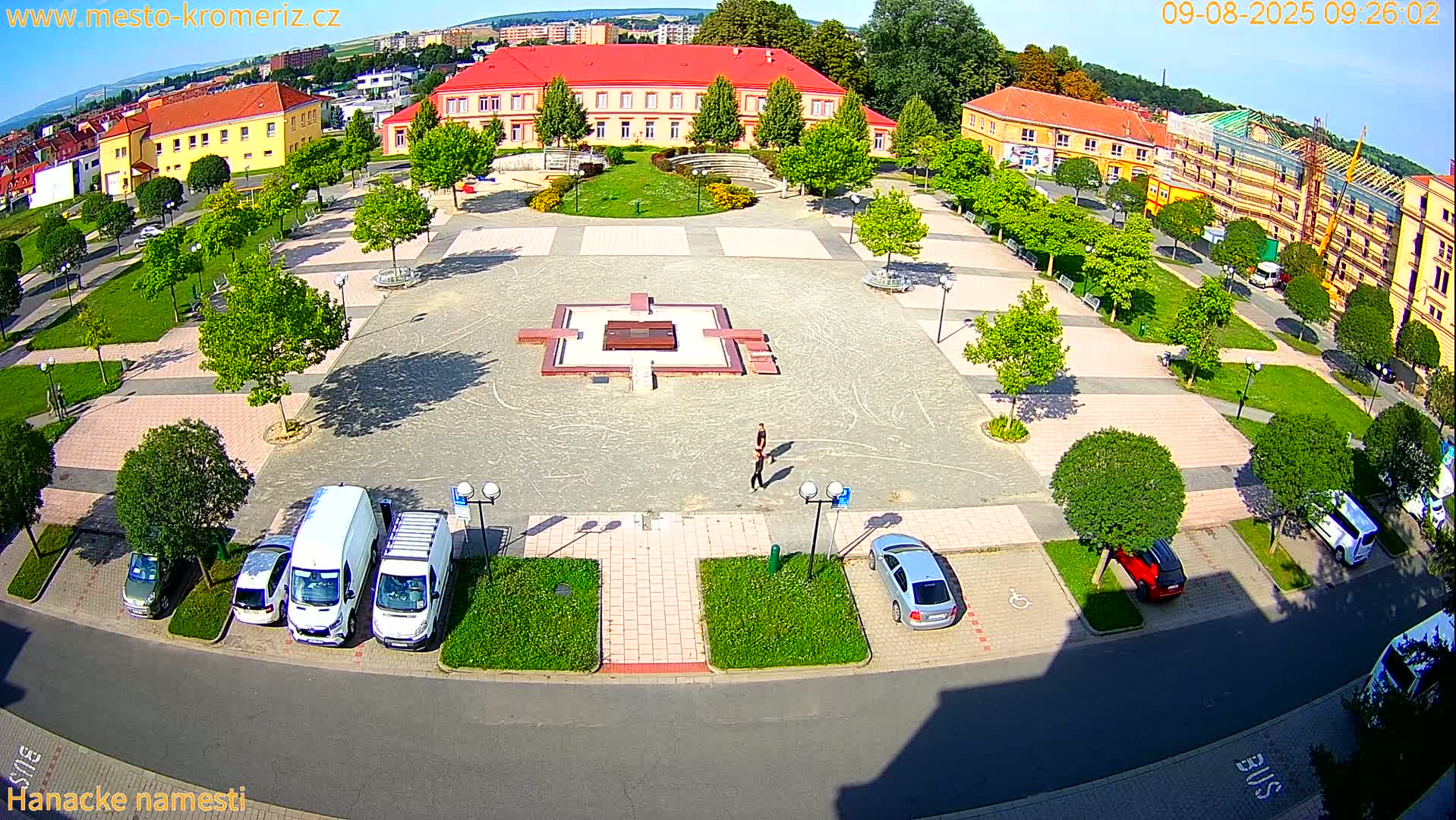 An aerial view shows a town square with a central fountain, surrounded by buildings, parked cars, and lush green trees under sunny conditions.