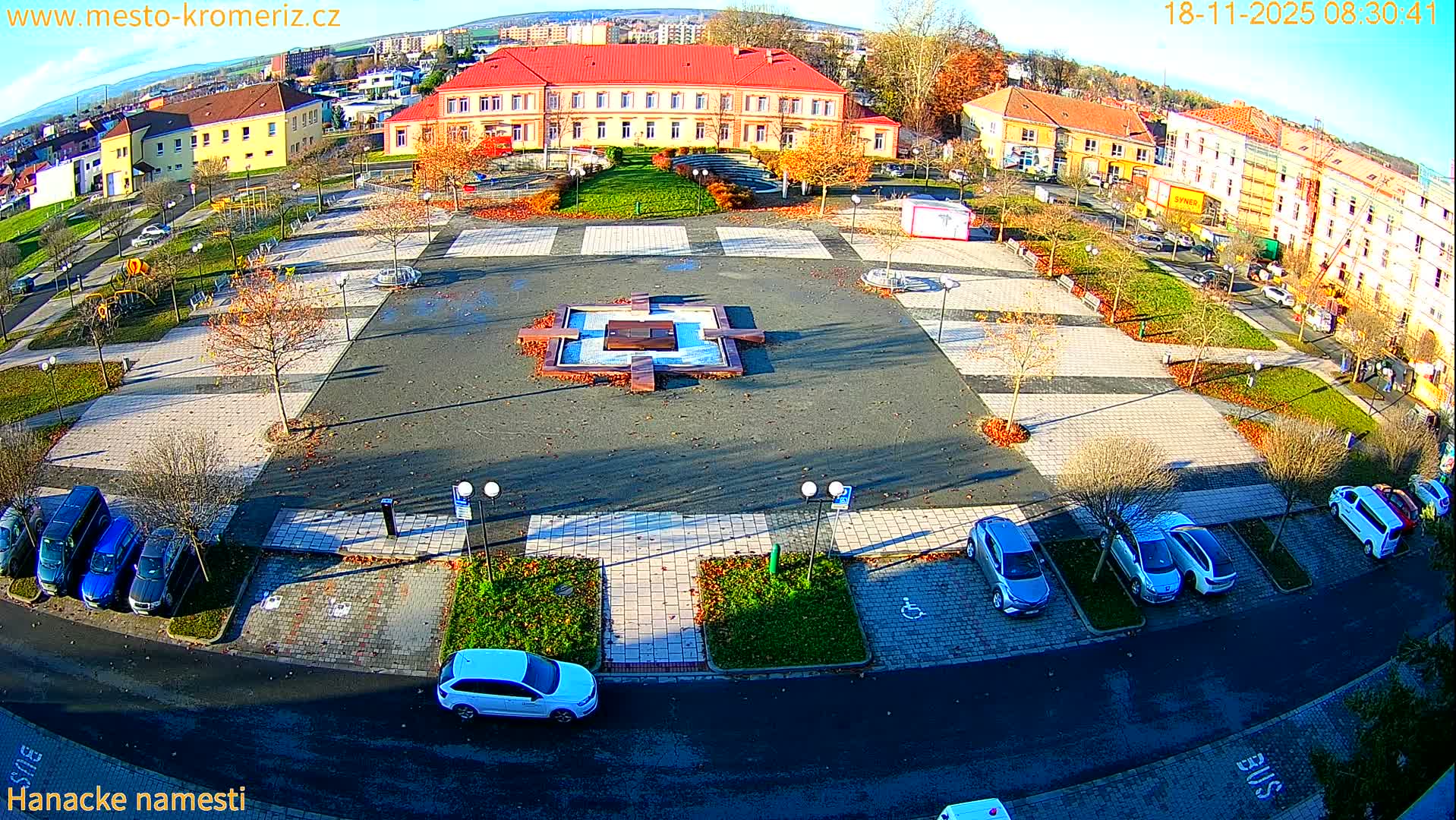 A sunny aerial view shows a town square with a central fountain, surrounding buildings, parked cars, and green landscaping.
