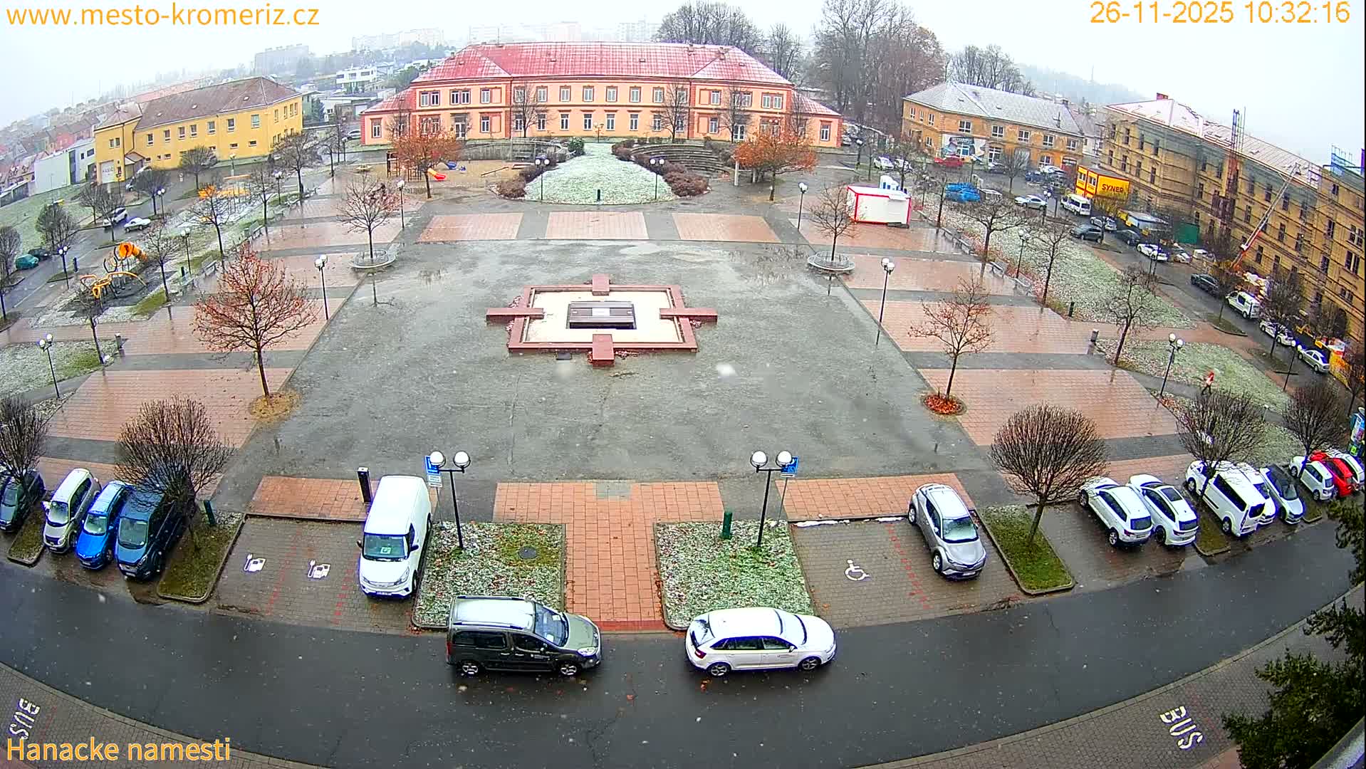 An overcast winter day reveals a wet town square with a light dusting of snow on grassy areas and bare trees, surrounded by various buildings and parked cars, with faint snow or sleet falling.