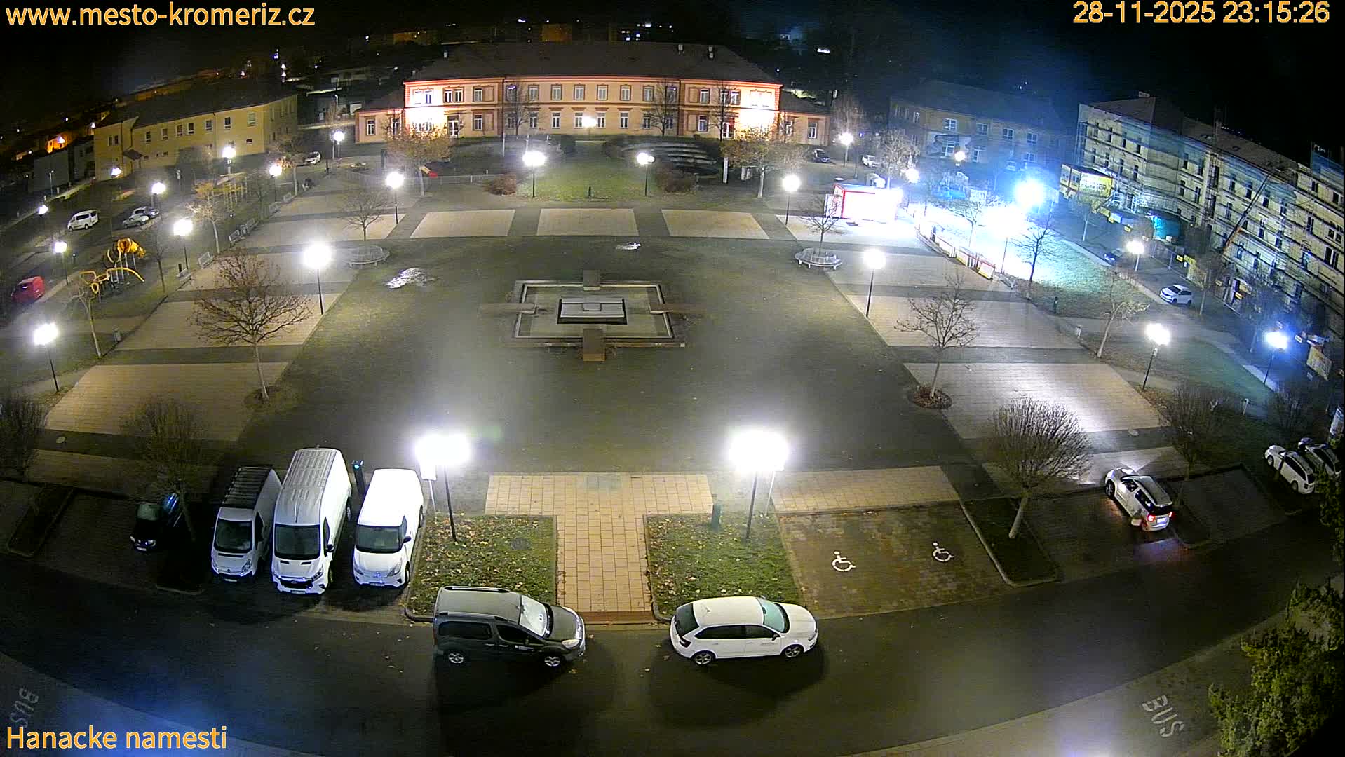 An overhead view captures a brightly lit town square at night, featuring a dry central fountain, bare trees, several parked vehicles, and surrounding buildings under a clear sky.