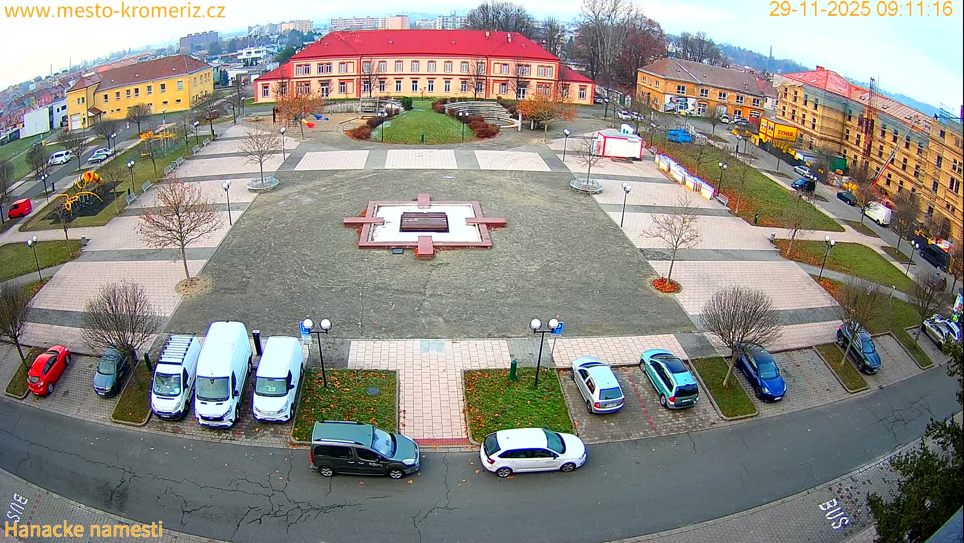 An elevated perspective reveals a town square with numerous parked cars, bare trees, and a central paved area featuring a monument, surrounded by buildings including a large red-roofed structure and one under construction, all beneath a bright, overcast sky.