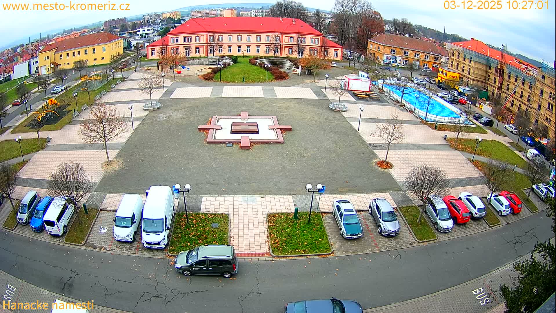 On an overcast late autumn or early winter day, an aerial perspective captures a quiet urban square featuring a central concrete monument, bare trees, numerous parked vehicles, a vibrant blue outdoor pool, and various buildings including a large red-roofed structure.