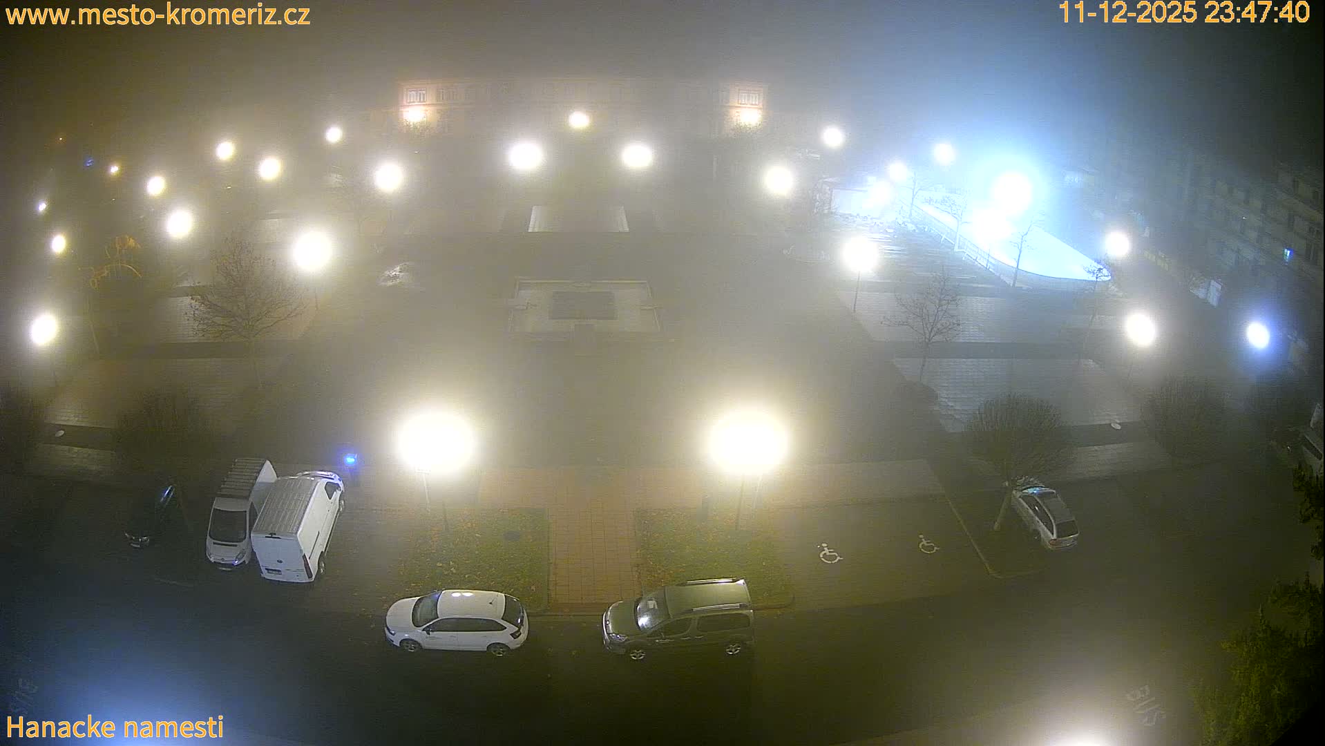 A foggy urban scene at night, viewed from above, features numerous streetlights illuminating parked cars, a public square, and surrounding buildings and bare trees.