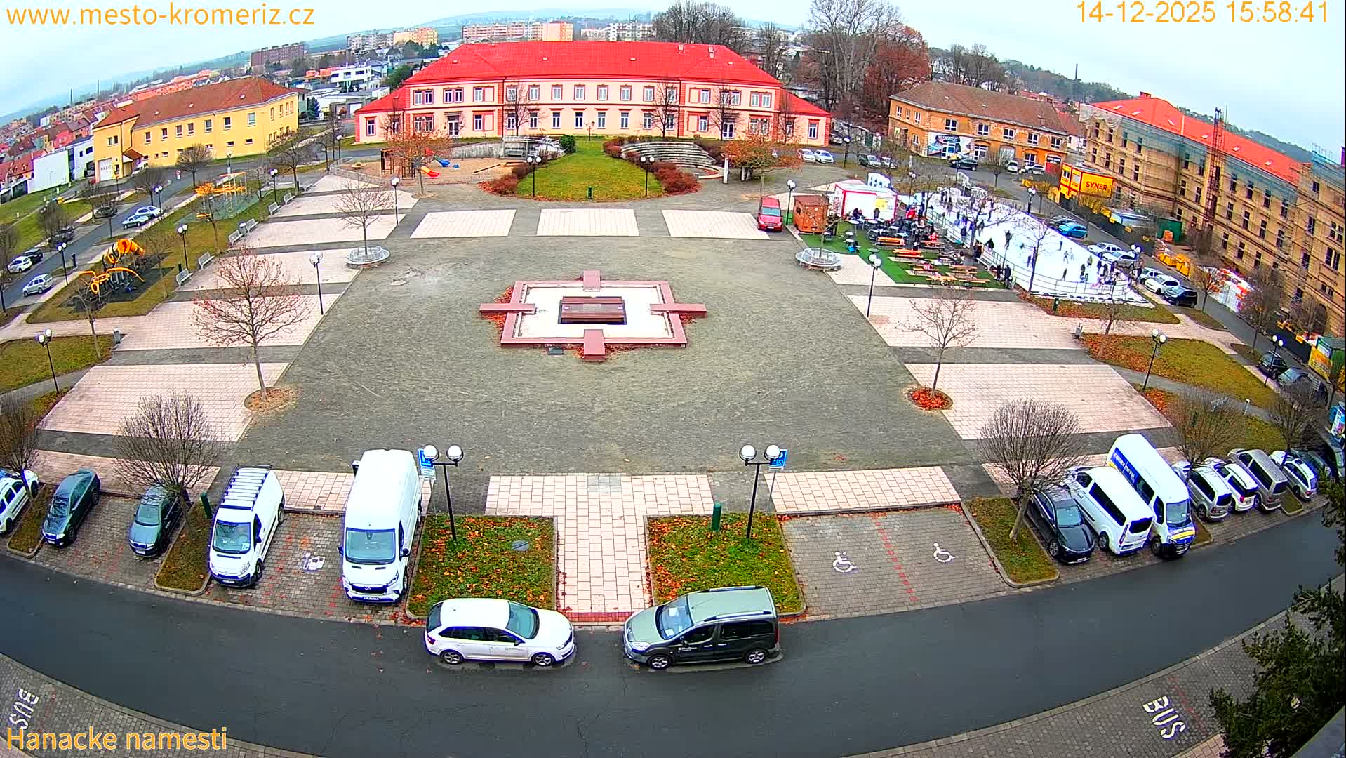 A foggy urban scene at night, viewed from above, features numerous streetlights illuminating parked cars, a public square, and surrounding buildings and bare trees.