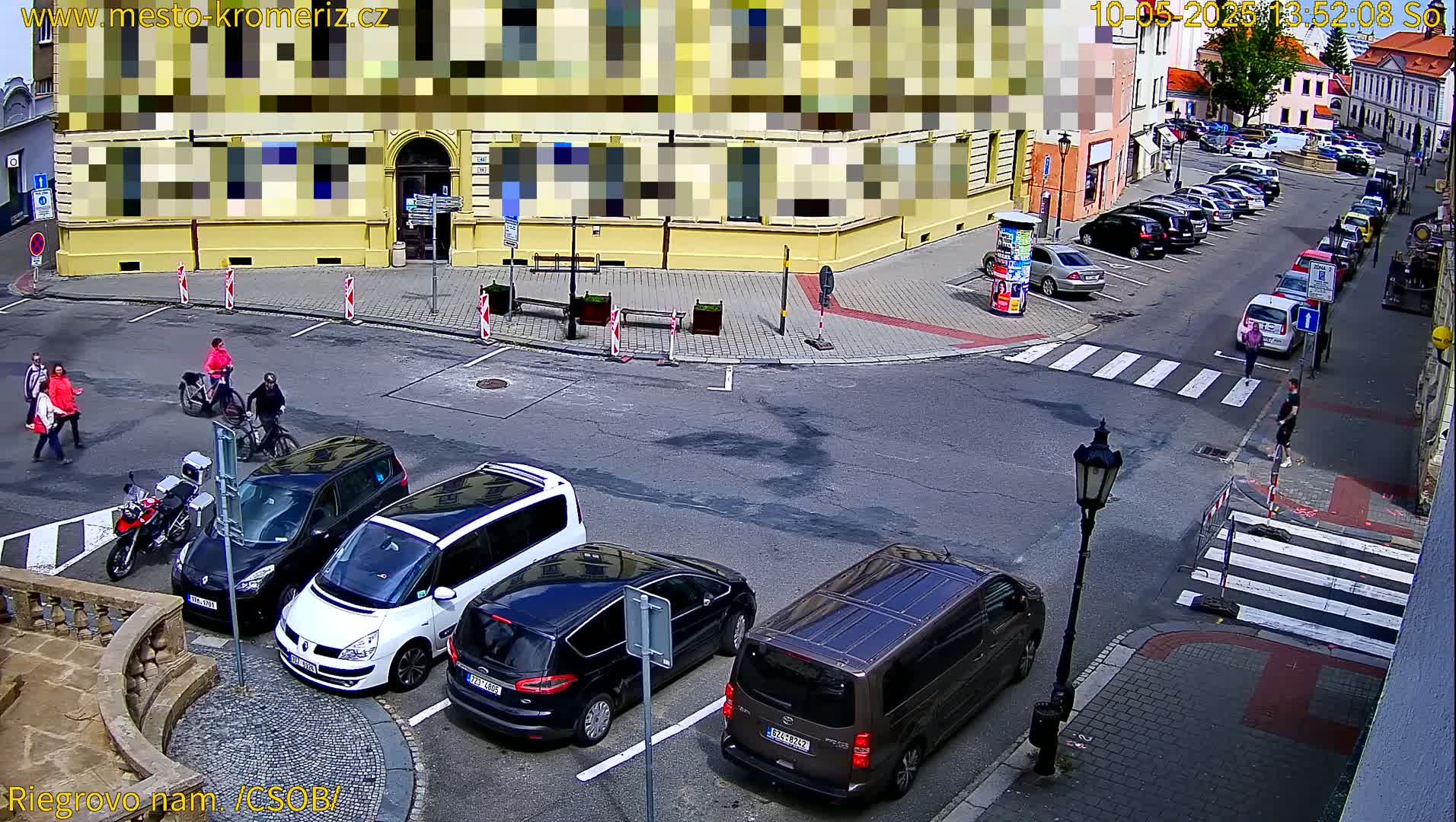 A sunny street corner scene shows several parked cars, pedestrians walking, and two cyclists riding past a yellow building.