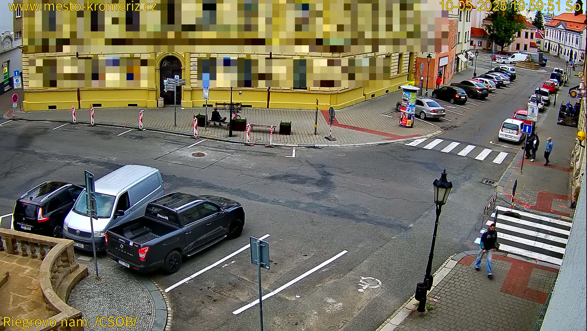A street corner with parked cars and a few pedestrians, under a partly sunny sky.