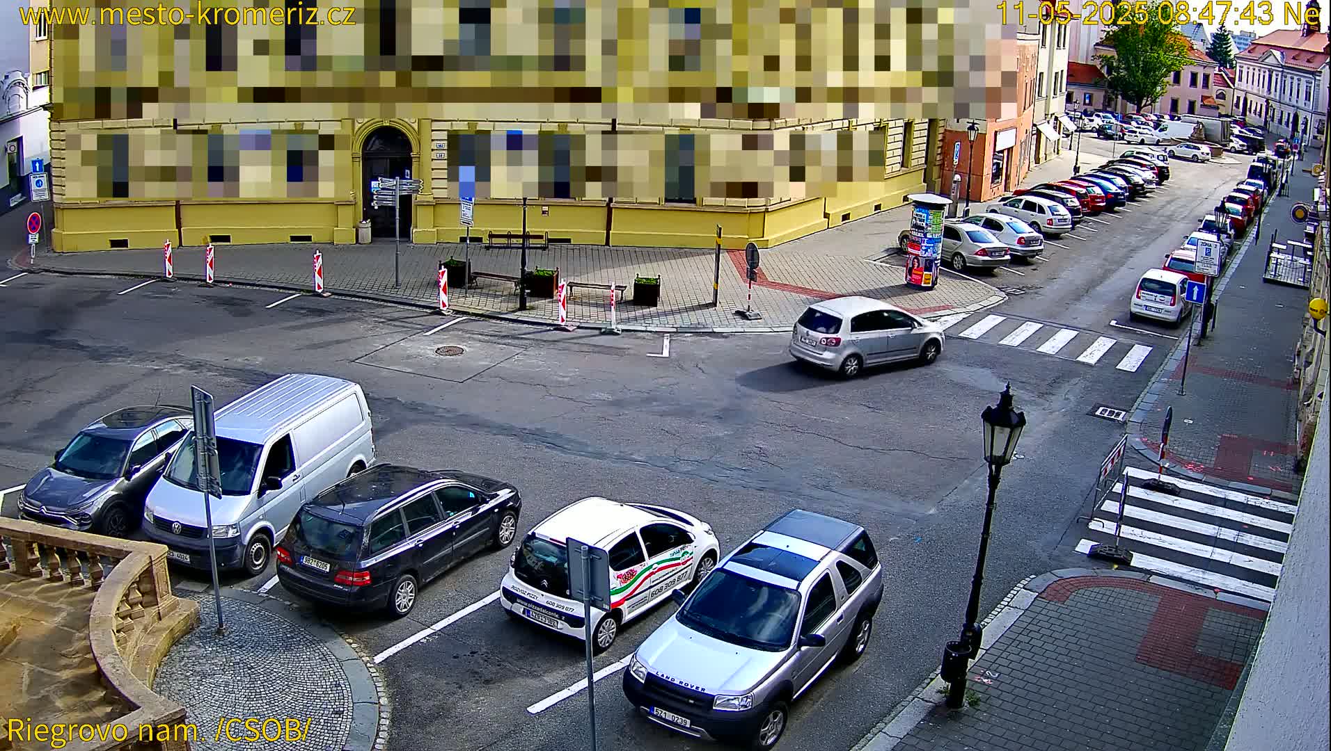 A street scene on a sunny day shows several cars parked along the curb and several others at a crossroads near a yellow building.
