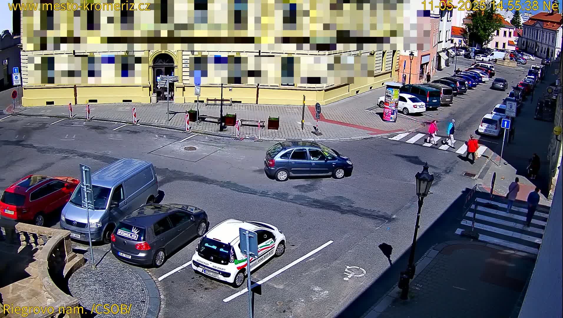 A sunny street scene shows several cars parked in a lot and on the street, with pedestrians crossing a crosswalk near a building.