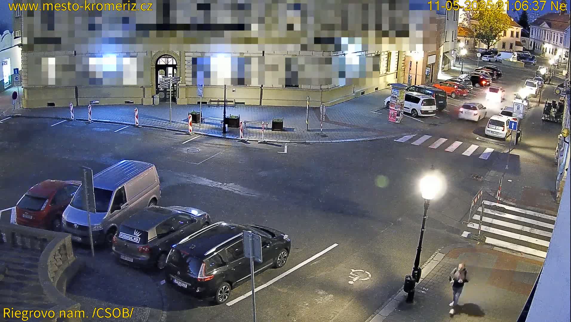 A nighttime, street-level view of a town square with several cars parked and one person walking, under artificial lighting.