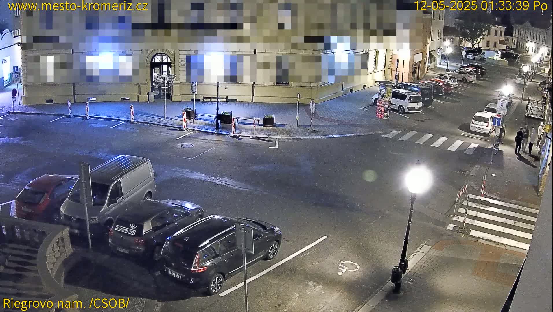 A nighttime, street-level view shows several cars parked along the curb of a street corner, with a few pedestrians visible near a crosswalk.