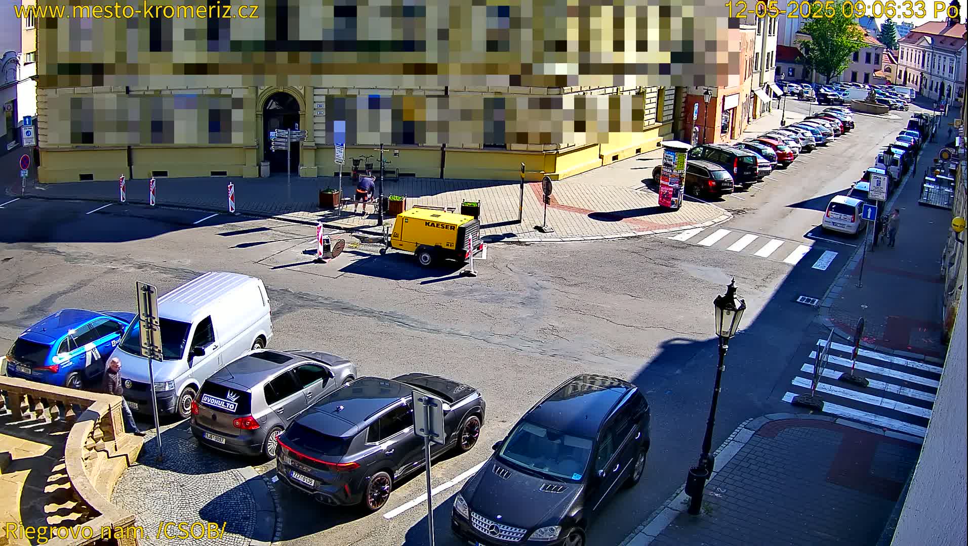 A sunny street scene shows several cars parked along the curb, a yellow generator, and a pedestrian crossing.