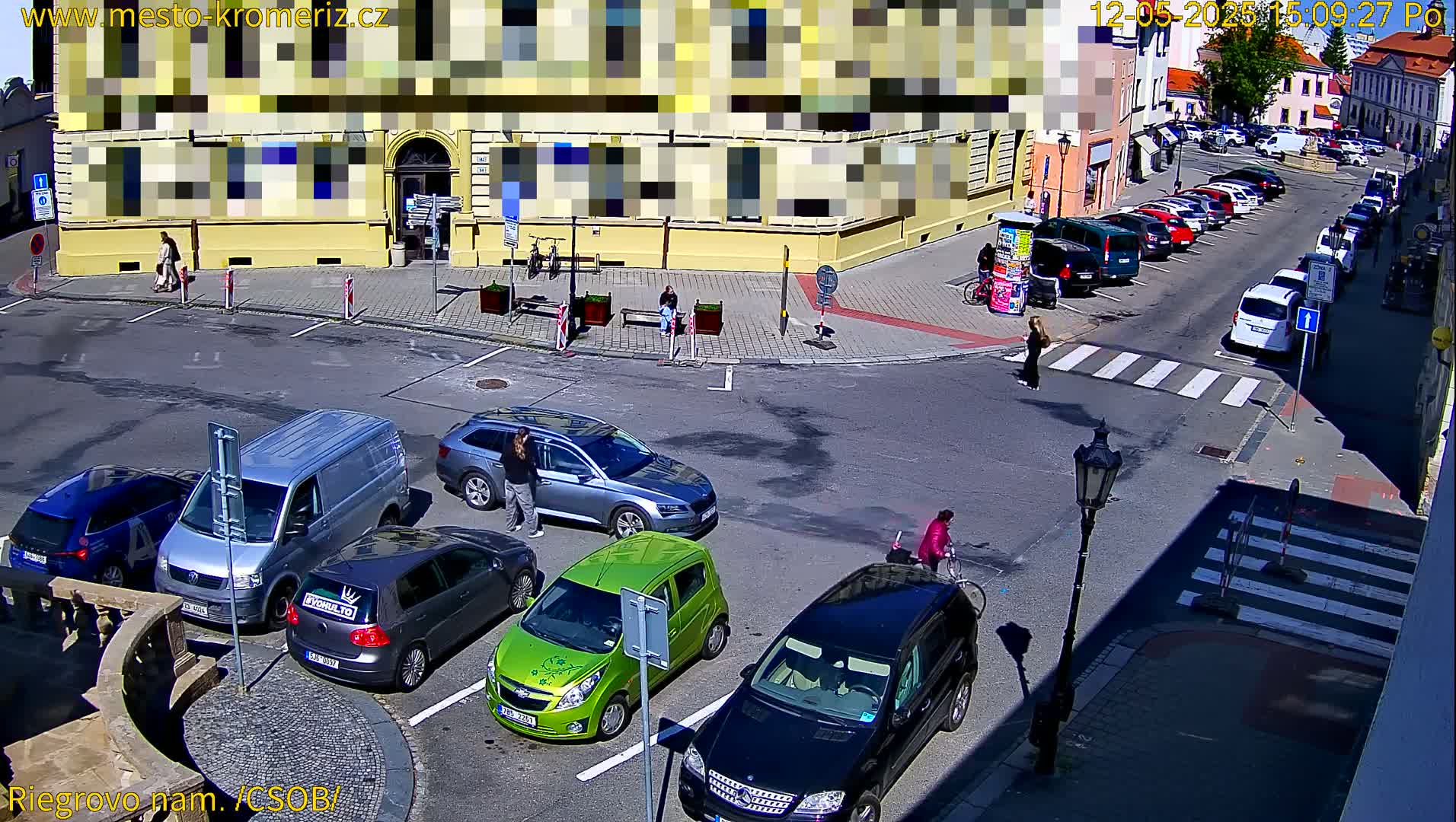 A sunny street scene shows several cars parked and driving around a town square with people walking and one person biking.
