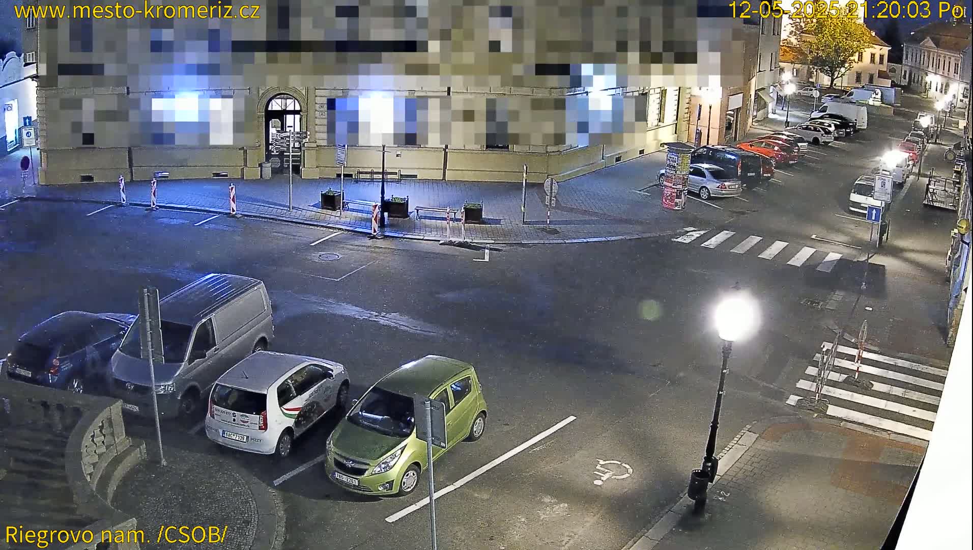 A nighttime street scene shows several cars parked in a lot and along the street near a building, under bright streetlights.