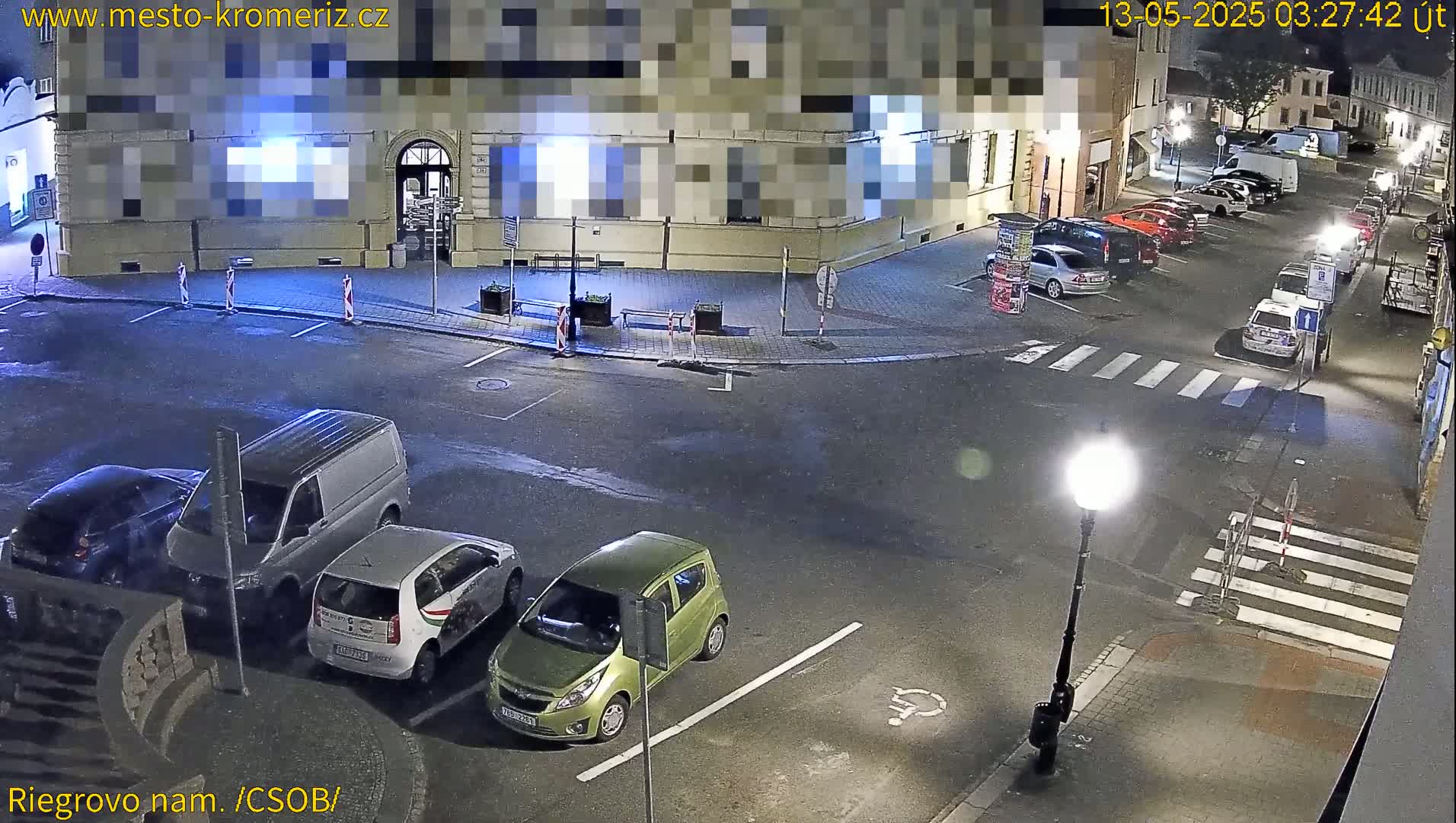 A nighttime street scene shows several cars parked along the sides of a street that intersects with another street, with a few cars parked in a small lot near a building.