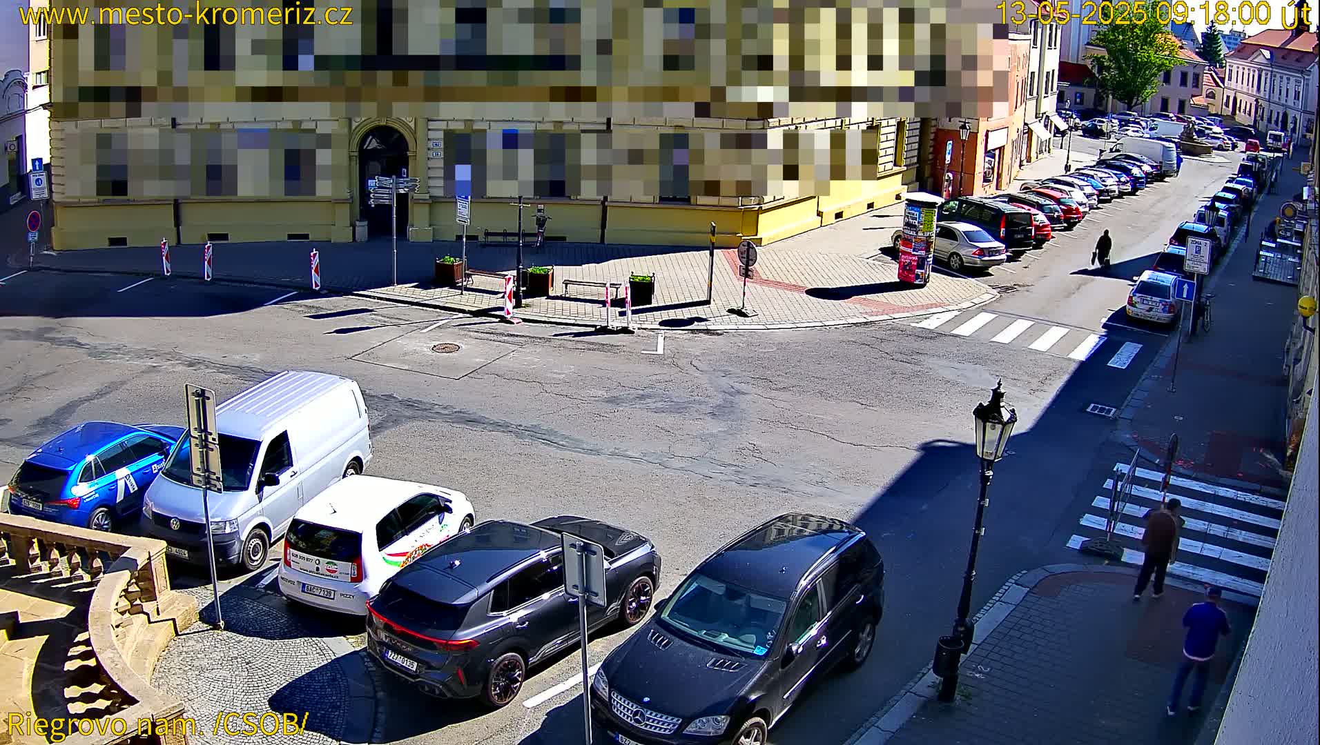 A sunny street corner shows several parked cars, a few pedestrians, and buildings lining the street.