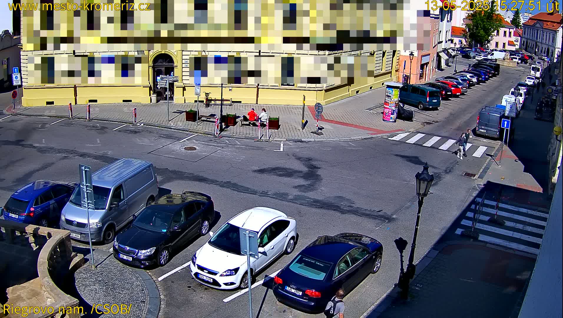 A sunny town square shows several parked cars, a van, and pedestrians crossing a crosswalk near a building.