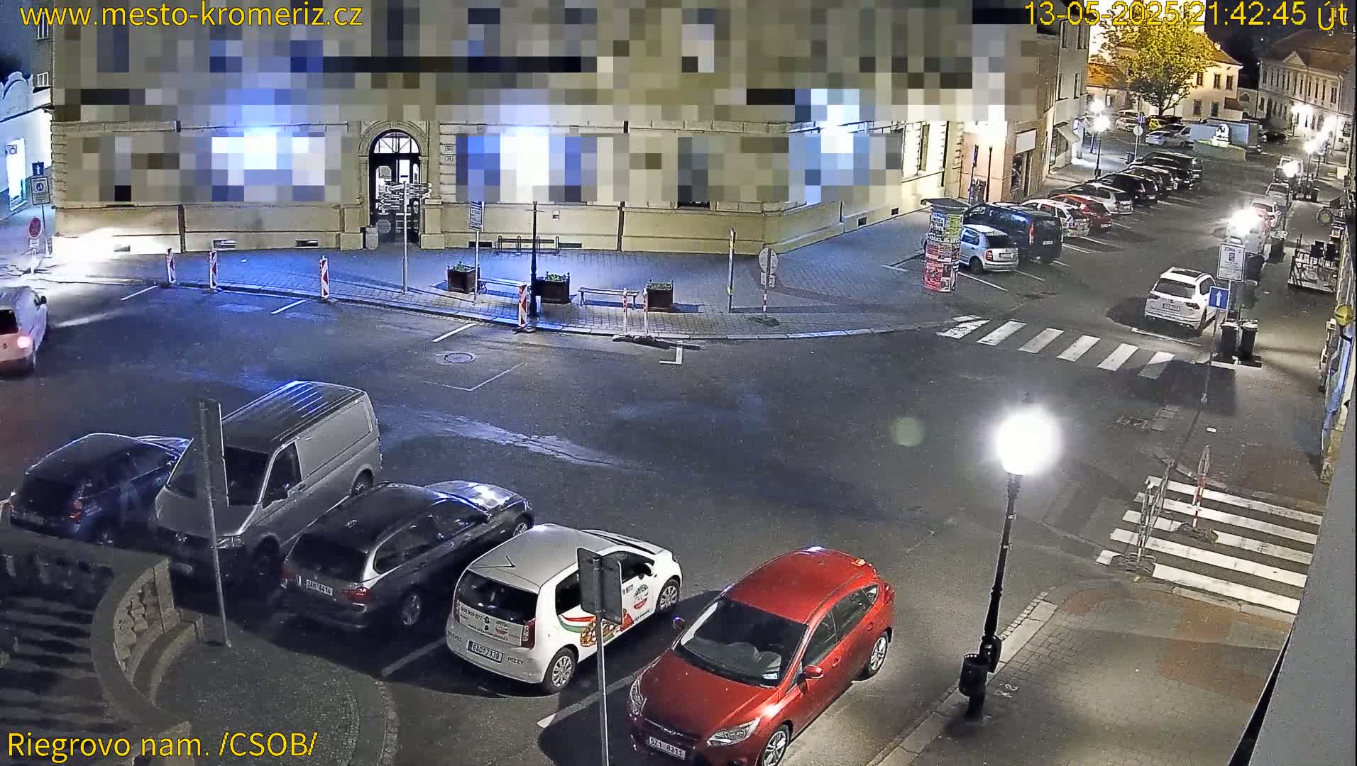 At night, several cars are parked in a town square near a building, with a few people walking around.