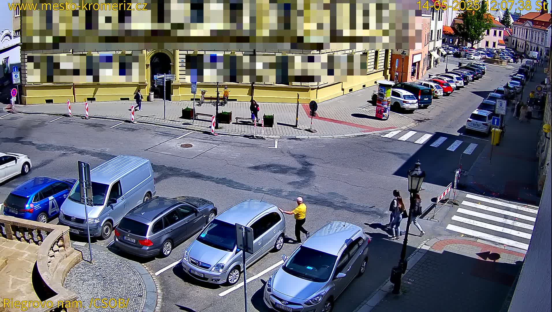A sunny street scene shows several parked cars, a van, and a few pedestrians near a building and a crosswalk.