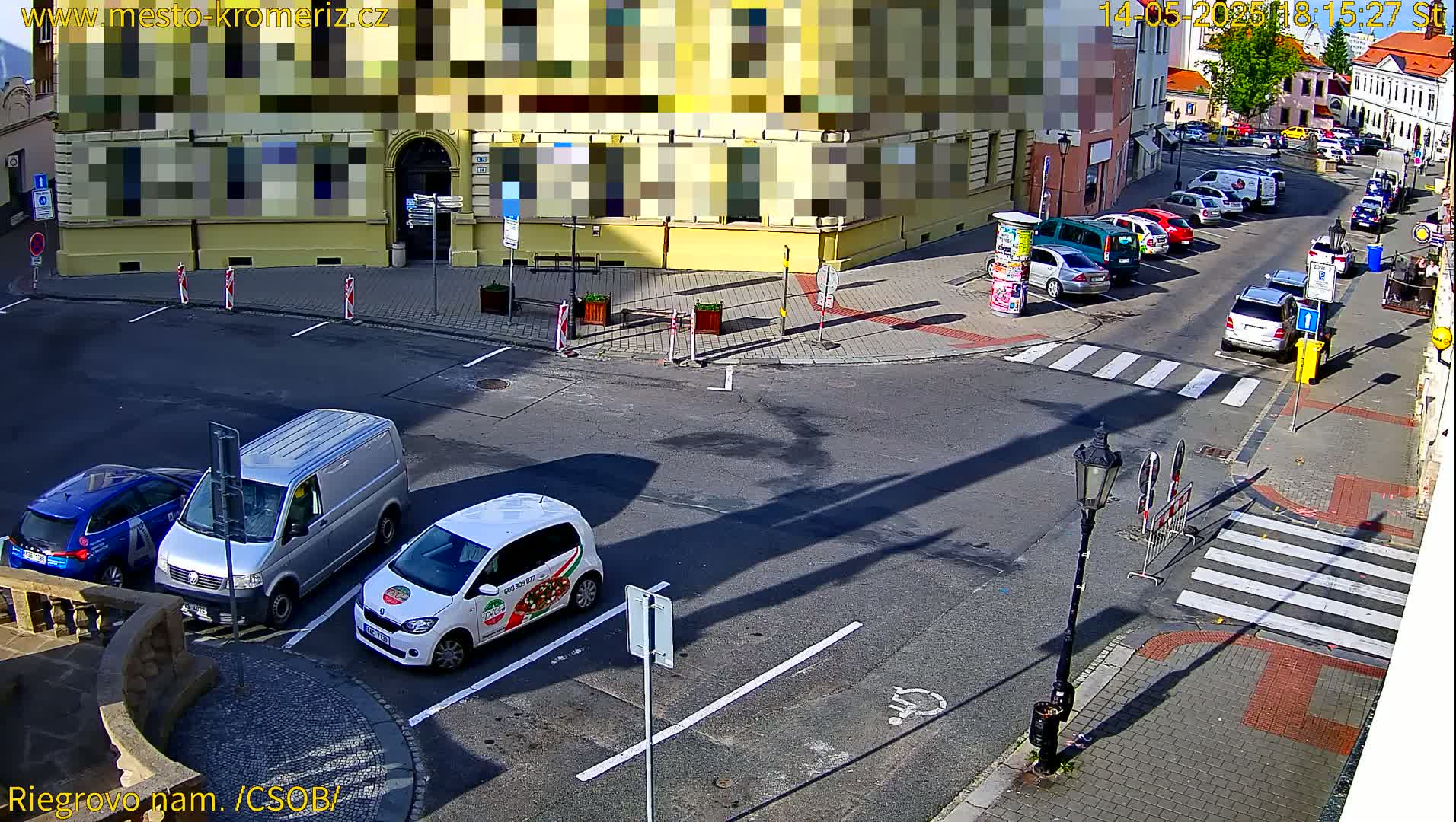 A sunny street scene shows several parked cars around a street corner, with a van and a pizza delivery car parked near the foreground.