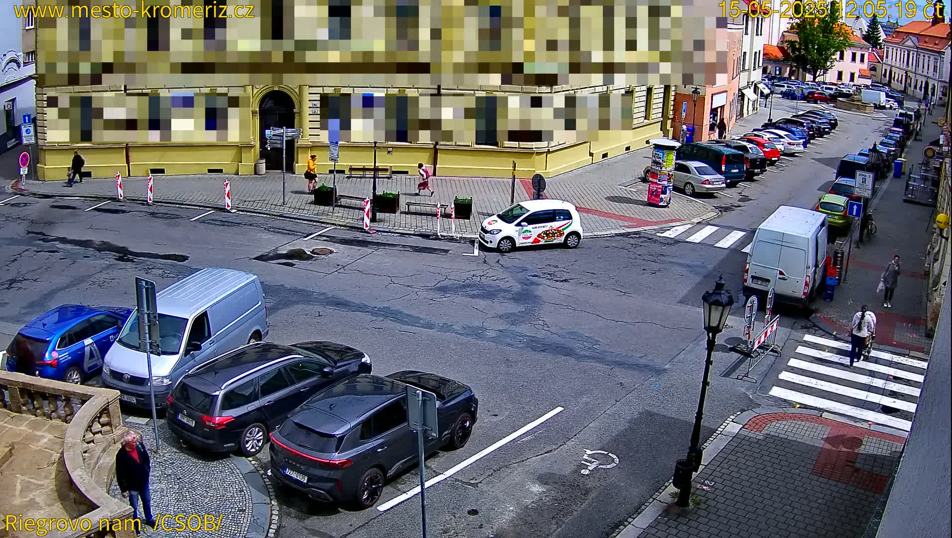A sunny street scene shows several cars parked along the curb, a few pedestrians walking, and some vehicles driving through a crossroads near a building.