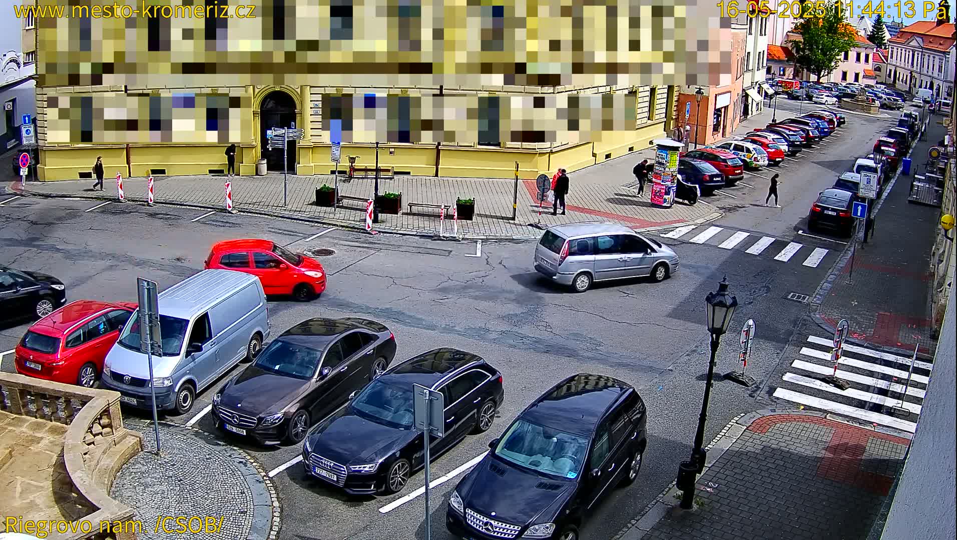 A sunny street scene shows several cars parked along the curb and several more in the street, with pedestrians walking on the sidewalk near a yellow building.
