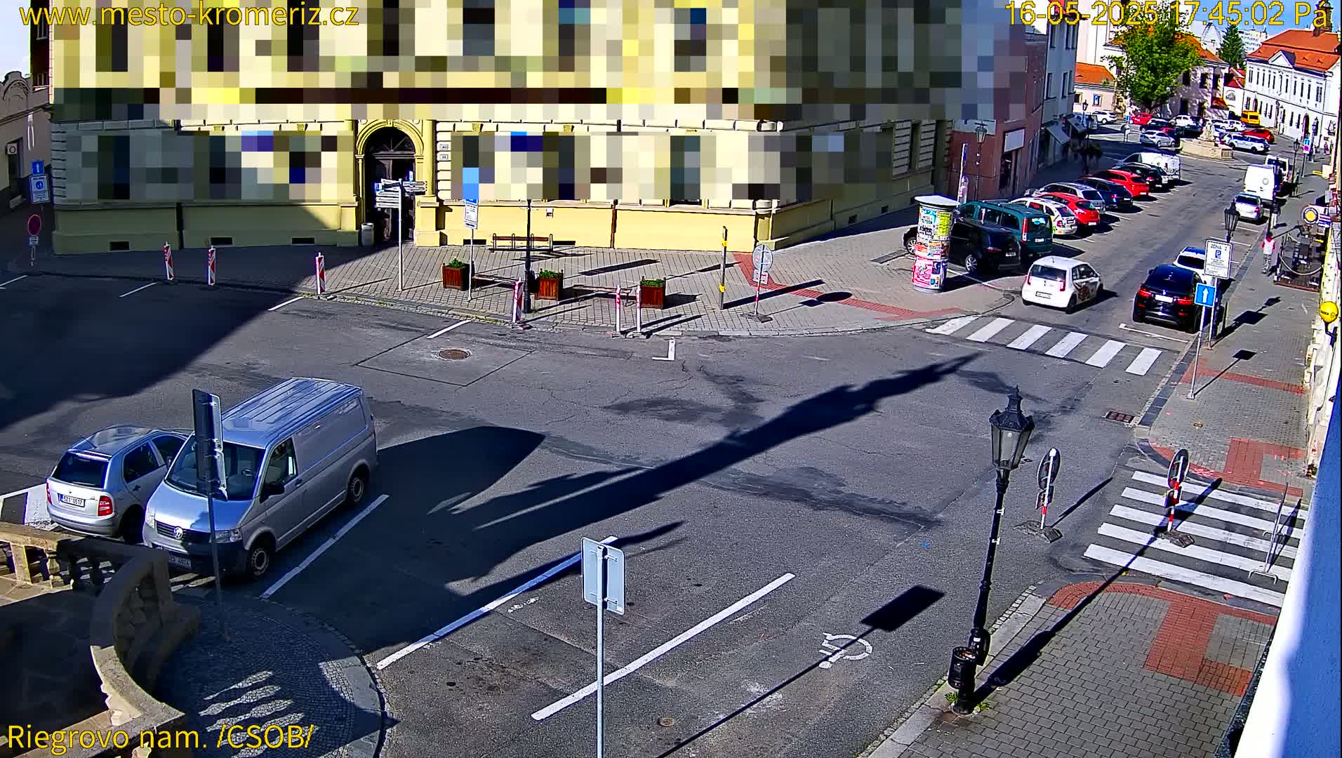 A sunny street scene shows a van and car parked near a building, with several other cars parked along a street and a crosswalk.
