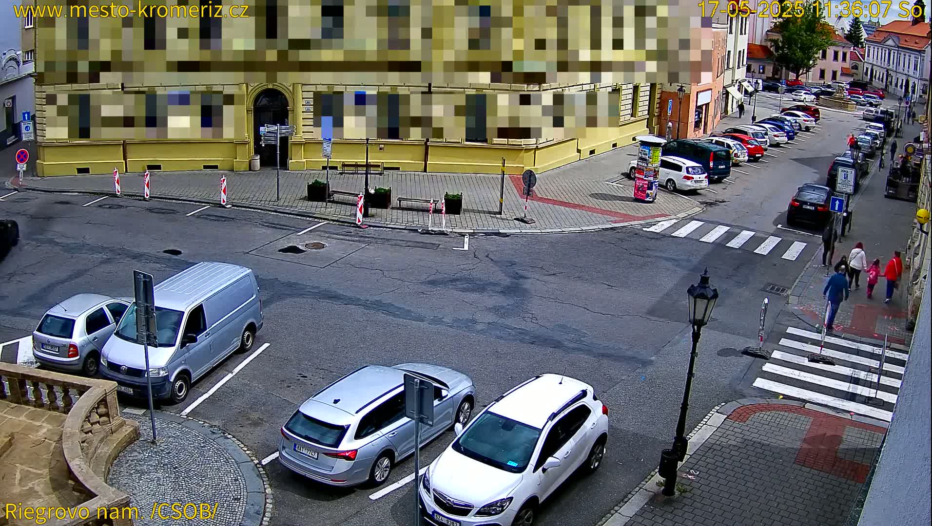 A street corner view shows several parked cars, a few pedestrians crossing a crosswalk, and a yellow building with its windows obscured.