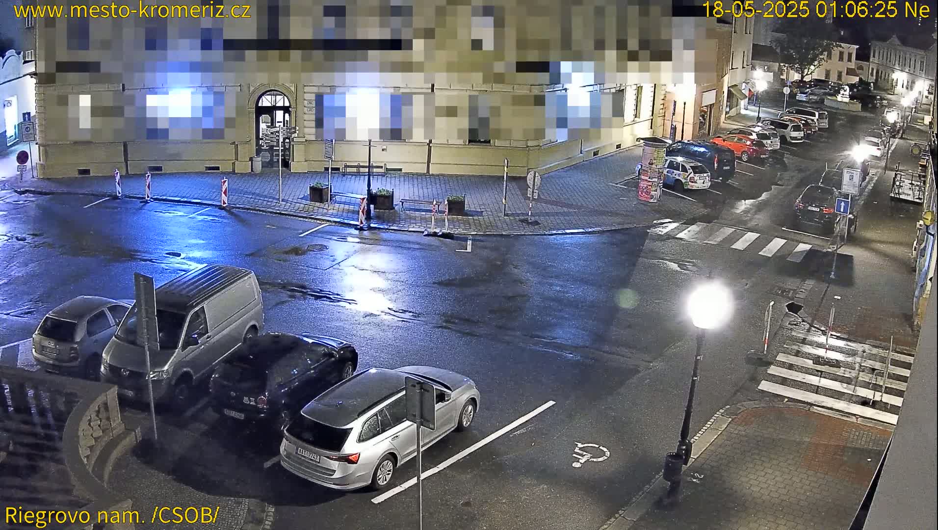 A wet nighttime street scene shows several cars parked in a lot and along the curb, with a few cars driving on a nearby road.