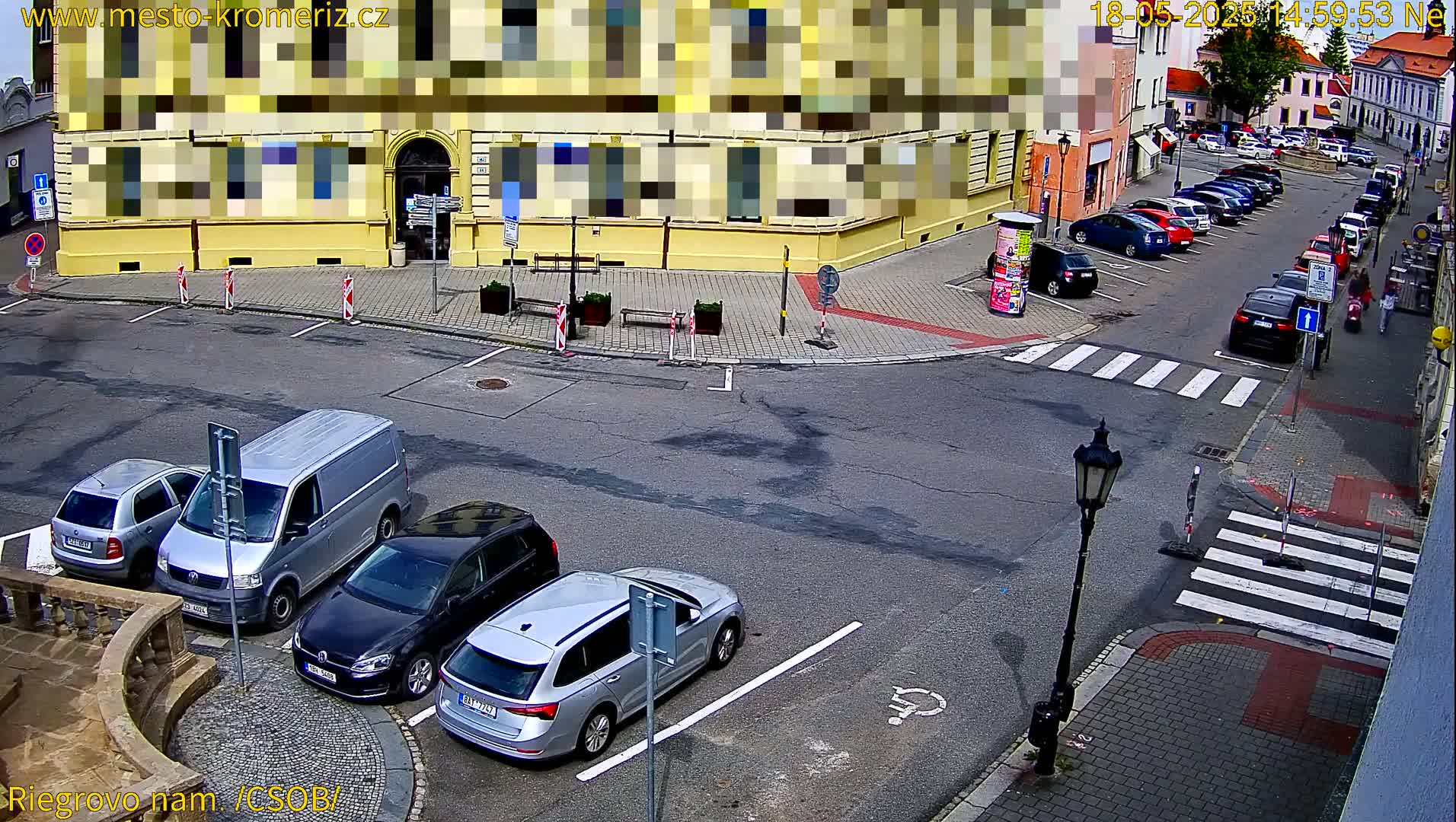 A sunny day shows a street corner with several parked cars, a van, and a building with its facade partially obscured.